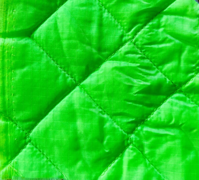 May include: Close-up of a vibrant green quilted fabric. The fabric features a diamond pattern formed by stitched lines. The material has a subtle sheen and a grid-like texture across its surface. The fabric's edges are visible on the left side.