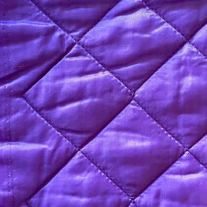 May include: Close-up of a quilted fabric in a vibrant purple hue. The fabric features a diamond pattern created by stitched lines. The texture appears smooth and slightly shiny, suggesting a synthetic material. The overall aesthetic is modern and clean.