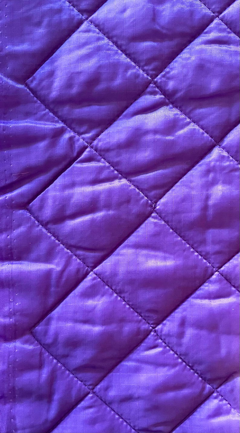 May include: Close-up of a quilted fabric in a vibrant purple hue. The fabric features a diamond pattern created by stitched lines. The texture appears smooth and slightly shiny, suggesting a synthetic material. The overall aesthetic is modern and clean.