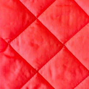 May include: Close-up of a vibrant red quilted fabric. The fabric is stitched in a diamond pattern, creating a textured surface. The material appears to be a lightweight, possibly synthetic, textile.