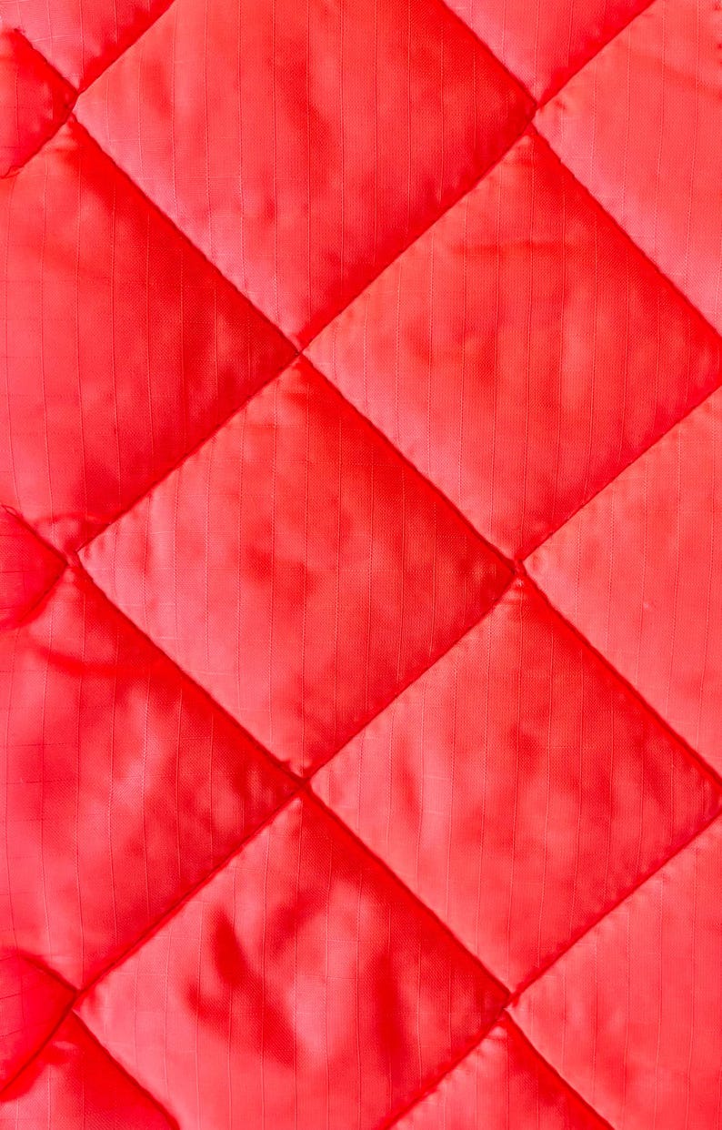 May include: Close-up of a vibrant red quilted fabric. The fabric is stitched in a diamond pattern, creating a textured surface. The material appears to be a lightweight, possibly synthetic, textile.