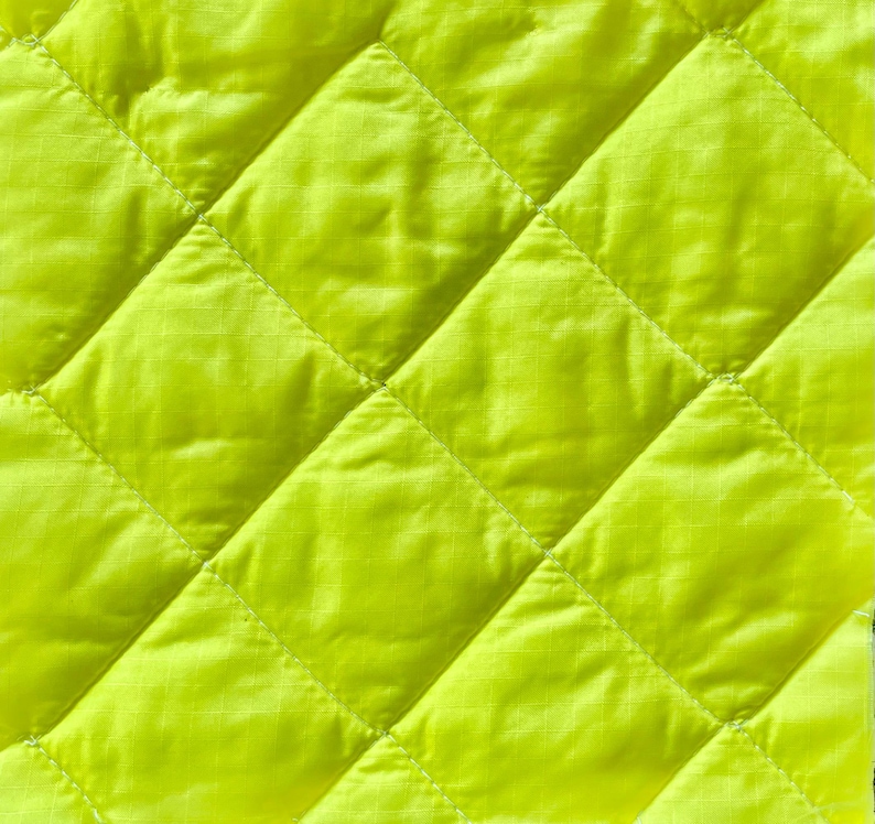 May include: Close-up of a bright yellow quilted fabric. The fabric features a diamond pattern created by stitched lines. The material appears smooth and slightly shiny, with subtle texture within each diamond.