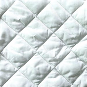 May include: Close-up of a white quilted fabric with a diamond pattern. The fabric has a slightly shiny appearance and is tightly stitched, creating a textured surface. The quilting is evenly spaced.