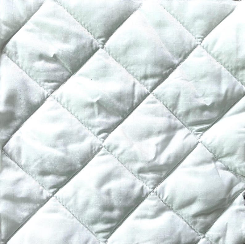 May include: Close-up of a white quilted fabric with a diamond pattern. The fabric has a slightly shiny appearance and is tightly stitched, creating a textured surface. The quilting is evenly spaced.
