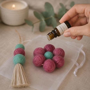 May include: A felted wool flower ornament with six magenta petals and a central mint green sphere, alongside a matching felted bead tassel. A small amber glass bottle labeled Lavender is positioned above the flower, dispensing oil.
