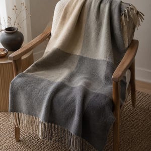 May include: A soft, woven throw blanket featuring a large-scale color-blocked pattern in shades of cream, light gray, and dark charcoal gray. The blanket has a textured surface and is finished with long, twisted fringe along the bottom edge.