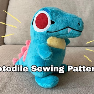 Pode incluir: Um brinquedo de pelúcia Totodile em tons de turquesa, rosa e amarelo, com um olho vermelho e branco. O texto "Totodile Sewing Pattern!" é exibido abaixo. O brinquedo está em um sofá de cor neutra.