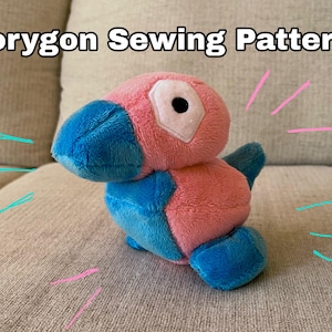 Könnte beinhalten: Ein rosa und blaues Porygon-Plüschtier mit einem weißen Auge und einer schwarzen Pupille. Der Text "Porygon Sewing Pattern!" steht oben. Das Spielzeug liegt auf einem beigen Sofa, mit rosa und blauen Linien darum.