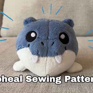 May include: A plush toy shaped like a blue and white seal with large black eyes and white fangs. The text "Spheal Sewing Pattern!" is displayed below the toy. Blue lines radiate from the toy.