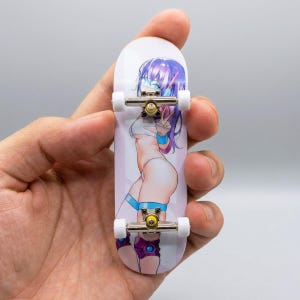 May include: A white fingerboard with a colorful anime-style illustration of a character with purple hair and blue accents. The miniature skateboard has silver metal trucks and white wheels. The fingerboard is held in a hand.