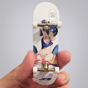 May include: A white fingerboard featuring a graphic of an anime-style character with blue hair, wearing a blue top and pink striped underwear. The board includes silver metal trucks, white wheels, and text in a speech bubble reading HOLD ON! GOSH, YOU ARE SO IMPATIENT. The JCS logo is printed at the bottom.