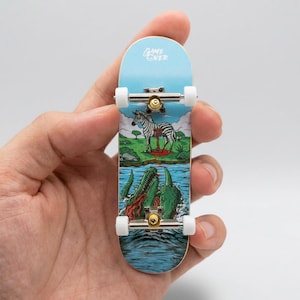 May include: A miniature skateboard with a blue top and a graphic design featuring a zebra and crocodiles. The skateboard has white wheels and metal axles. The top of the board has the words "Game Over".