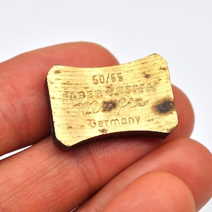 Op de afbeelding: Een klein, rechthoekig, goudkleurig metalen object met afgeronde randen. De tekst "50/65", "FABER-CASTELL", "Mifix" en "Germany" zijn in het oppervlak geëtst. Het object heeft een versleten, vintage uitstraling.