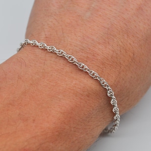 May include: A silver rope chain bracelet. The bracelet is made of small, intertwined links, creating a textured appearance. The jewelry is worn on a wrist.