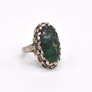 May include: A silver ring with an oval, green gemstone with dark markings. The stone is set in a decorative bezel with a leaf design. The ring has a detailed band and a vintage style.