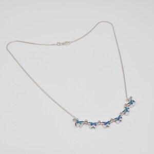 Op de afbeelding: Een zilverkleurige ketting met een reeks kleine, vlindervormige bedels aan de onderkant. Elke vlinderbedel is ingelegd met iriserend blauw en groen opaalachtig materiaal, verbonden door dunne zilverkleurige metalen schakels.