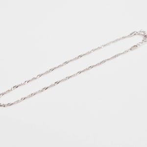 May include: A silver-toned chain necklace featuring a twisted, shimmering link design. The necklace includes a small metal clasp mechanism and a tiny heart-shaped charm attached near the end of the chain, creating a reflective surface.