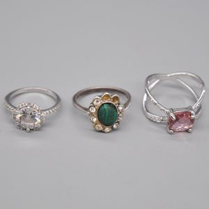 Puede incluir: Tres anillos de plata con piedras preciosas de diferentes colores. El primer anillo tiene una piedra redonda transparente, el segundo anillo tiene una piedra ovalada verde y el tercer anillo tiene una piedra ovalada rosa.