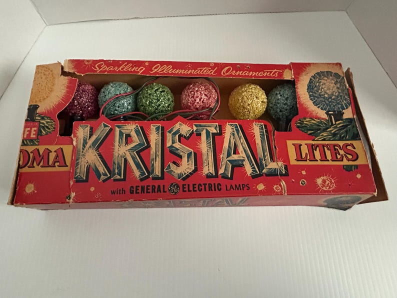 May include: A box of vintage Christmas lights with the brand name "Kristal Lites" and the text "Sparkling Illuminated Ornaments with General Electric Lamps". The box contains a string of multi-colored, sparkly, round Christmas lights.