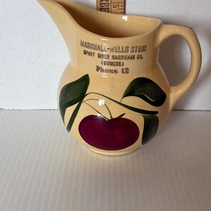 Puede incluir: Jarra de cerámica color crema con asa y boquilla. La jarra presenta un diseño pintado de una gran cereza roja oscura con hojas verdes. El texto "MARSHALL-WELLS STORE" y otro texto está impreso en la jarra.