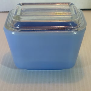 Op de afbeelding: Een vierkante, lichtblauwe glazen container met een doorzichtig glazen deksel. De container heeft een licht taps toelopende vorm en een gladde, matte afwerking. Het deksel is rechthoekig met horizontale ribbels.