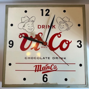 Puede incluir: Un reloj vintage con una esfera blanca y agujas negras. El reloj anuncia una bebida de chocolate llamada "Vi Co" con el lema "Drink Vi Co Chocolate Drink". El reloj también tiene el texto "ManCo" y "Reg'd."