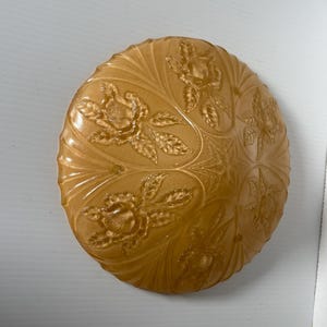 Może przedstawiać: Okrągły, bursztynowy szklany talerz z kwiatowym wzorem róż. Talerz ma wypukły, wytłoczony wzór róż i liści. Talerz jest prawdopodobnie zabytkowy lub antyczny i jest przedmiotem dekoracyjnym.