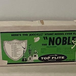 May include: A white cardboard box with a green and white label. The label features a black and white illustration of a plane with the text "HERE'S THE MIRACULOUS STUNT MODEL, EVEN FLEW WHEN" and "THE "NOBLER" TOP FLITE" printed on it.