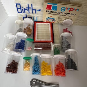 Könnte beinhalten: Ein Vintage Snapics Transportation Set mit Kunststofffliesen in verschiedenen Farben, einem Rahmen und einem Werkzeug. Das Set enthält eine weiße Kunststoffleinwand mit dem Wort "Birth-" in Blau. Auf der Box steht "Kunststofffliesen, die Bilder im Handumdrehen machen!"