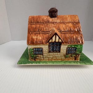 Puede incluir: Un recipiente de cerámica con forma de cabaña con tejado de paja y chimenea, sobre una base rectangular verde. La cabaña presenta una fachada de piedra, ventanas y vegetación decorativa. El tejado es marrón y la chimenea es marrón oscuro.