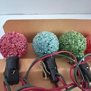 May include: A string of Christmas lights with four bulbs. The bulbs are round and have a textured surface. The bulbs are pink, blue, green, and pink. The lights are wired together with black wires.