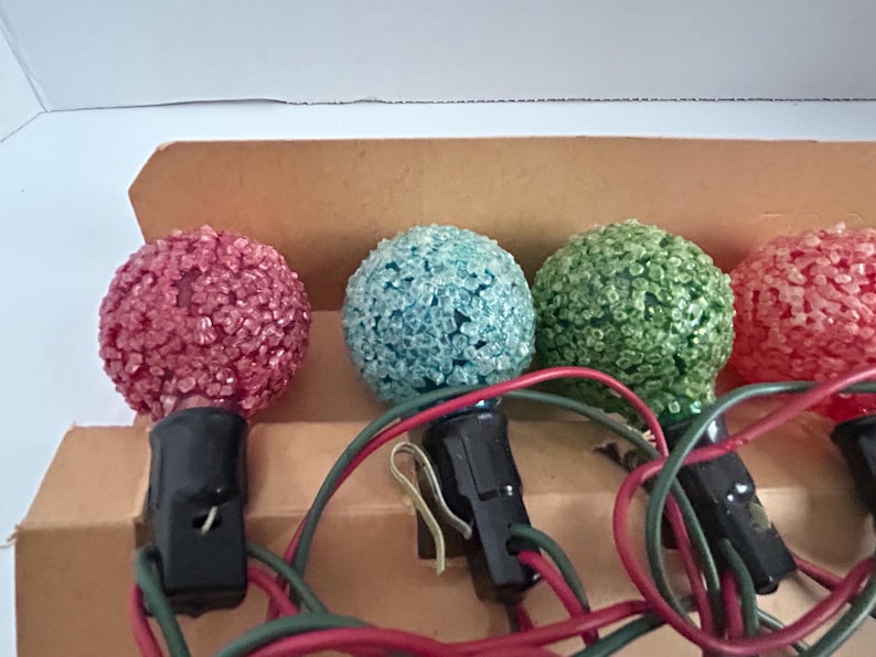 May include: A string of Christmas lights with four bulbs. The bulbs are round and have a textured surface. The bulbs are pink, blue, green, and pink. The lights are wired together with black wires.