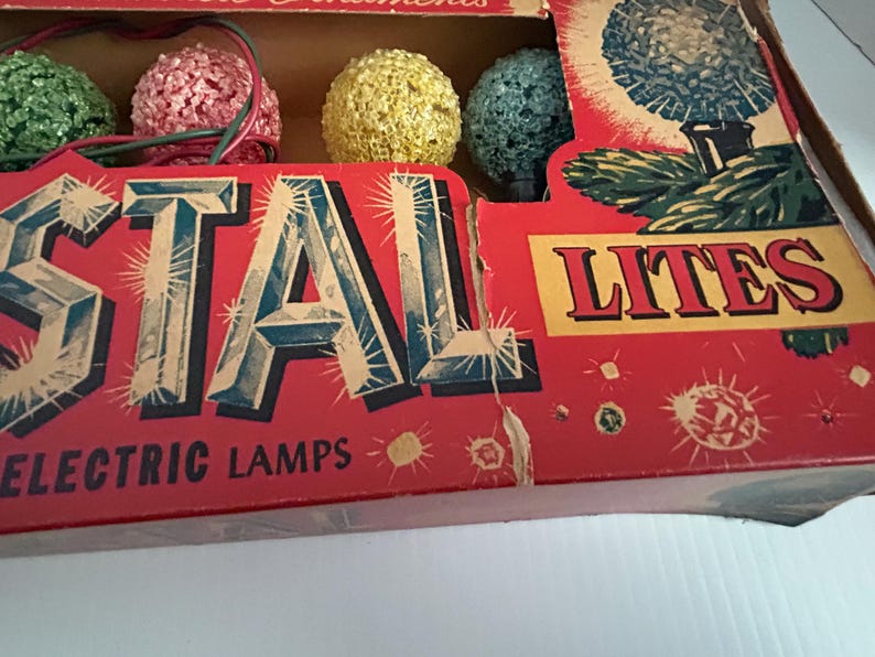 May include: Vintage box of Christmas lights with the words "Crystal Lites Electric Lamps" on the front. The box features a red background with a white border and a variety of colorful lights.
