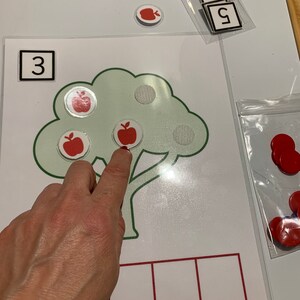 May include: A green tree with a brown trunk and three red apples on it. The number 3 is written in a black square at the top of the image. There are five empty squares at the bottom of the image.
