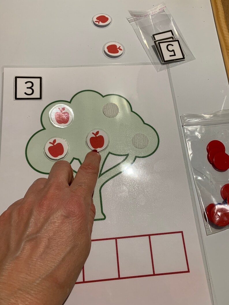 May include: A green tree with a brown trunk and three red apples on it. The number 3 is written in a black square at the top of the image. There are five empty squares at the bottom of the image.