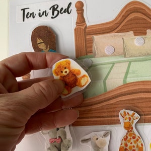 May include: A felt board game with the title "Ten in Bed" printed on it. The board features a bed with a brown headboard and a green and white patterned bedspread. There are several felt animal figures, including a giraffe, a donkey, an elephant, and a teddy bear.