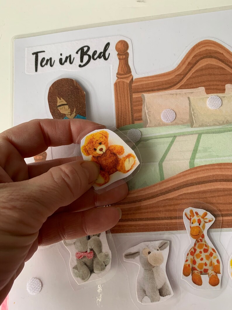 May include: A felt board game with the title "Ten in Bed" printed on it. The board features a bed with a brown headboard and a green and white patterned bedspread. There are several felt animal figures, including a giraffe, a donkey, an elephant, and a teddy bear.