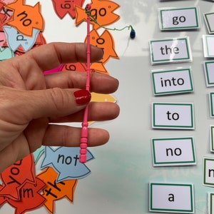 May include: A pink plastic fishing rod with a green string and a blue bobber. The rod is being held by a hand. There are several colourful fish-shaped cards with words on them, including "go", "not", "the", "into", "to", "no", and "a".