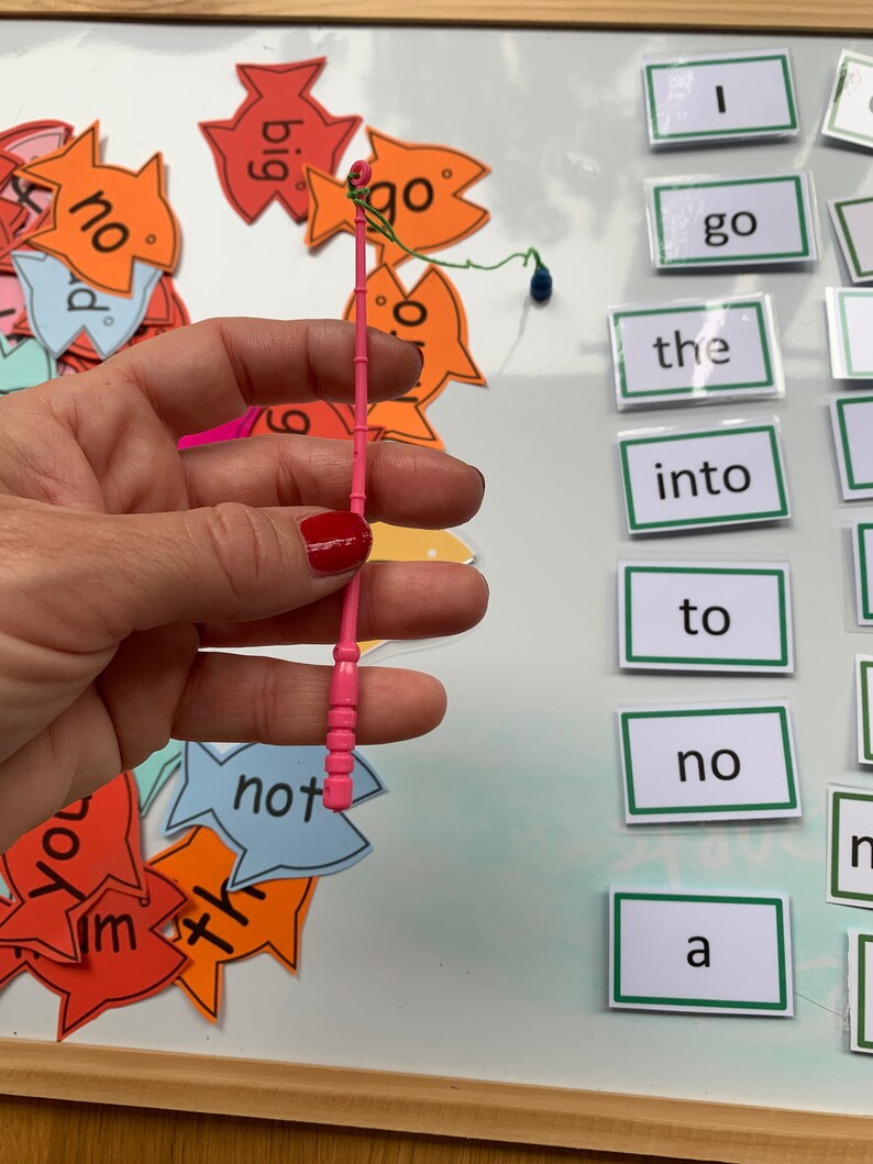 May include: A pink plastic fishing rod with a green string and a blue bobber. The rod is being held by a hand. There are several colourful fish-shaped cards with words on them, including "go", "not", "the", "into", "to", "no", and "a".
