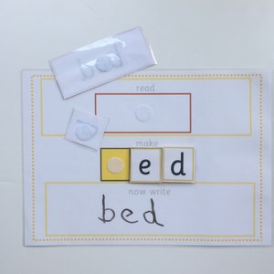 May include: A white card with the word "bed" written on it. The word is made up of three letter tiles, one yellow with a circle, one white with the letter "e" and one white with the letter "d". The word "bed" is written below the tiles. The card also has the words "read", "make", and "now write" printed on it.