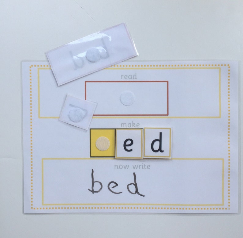 May include: A white card with the word "bed" written on it. The word is made up of three letter tiles, one yellow with a circle, one white with the letter "e" and one white with the letter "d". The word "bed" is written below the tiles. The card also has the words "read", "make", and "now write" printed on it.