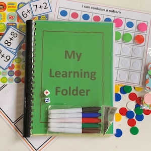 May include: A green binder with the words "My Learning Folder" on the cover. The binder is surrounded by various educational materials, including colourful stickers, number flashcards, a dice, dry-erase markers, and a pattern recognition worksheet.