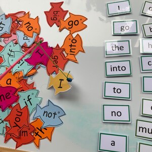 May include: A white board with a fishing game for learning basic English words. The game includes colourful fish with words like "go", "not", "big", "get", and "I".  The words are on a white background with black lettering.  The board also has a set of word cards with words like "the", "into", "to", "no", and "a".