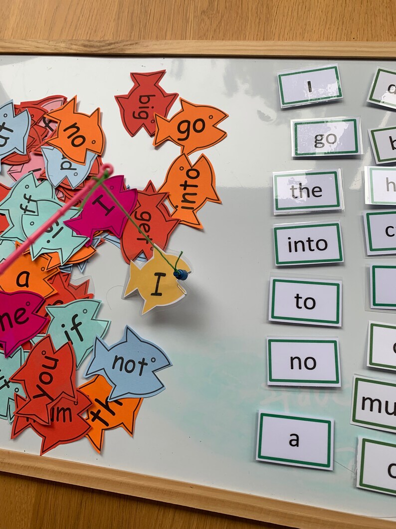 May include: A white board with a fishing game for learning basic English words. The game includes colourful fish with words like "go", "not", "big", "get", and "I".  The words are on a white background with black lettering.  The board also has a set of word cards with words like "the", "into", "to", "no", and "a".
