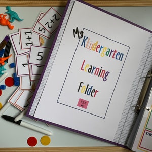 May include: A purple binder with the text "My Kindergarten Learning Folder" on the front. The binder is open to reveal a page with the text "Tuesday Maths" and "Wednesday". There are also colourful dinosaur toys, markers, and number flashcards on a white surface.
