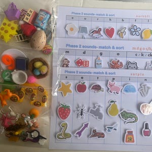 May include: A selection of miniature toys and educational resources in a clear bag. Includes small items such as numbers, a nest with eggs, and a tambourine. Also features printed sheets with the text "Phase 2 sounds-match & sort".