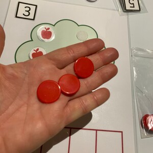 May include: A hand holding four red counting chips. The chips are on a white surface with a green cloud shape and a number 3 in a white square. There are also red apple stickers on the surface.
