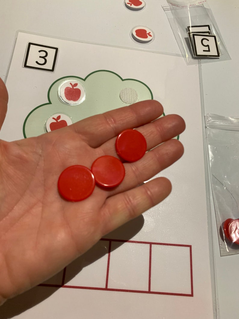 May include: A hand holding four red counting chips. The chips are on a white surface with a green cloud shape and a number 3 in a white square. There are also red apple stickers on the surface.