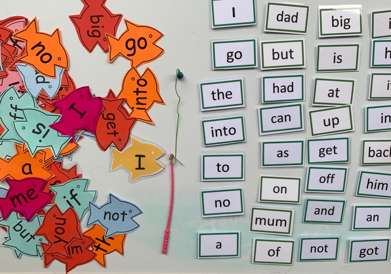 May include: A set of colourful fish-shaped cards with words printed on them, such as "go", "big", "not", "I", and "get".  The cards are arranged in a pile on a white surface.  There is a green and pink string attached to a blue bead, and a set of white cards with green borders, each with a single word printed on them, such as "dad", "the", "into", "to", "no", "mum", "of", "not", and "got".