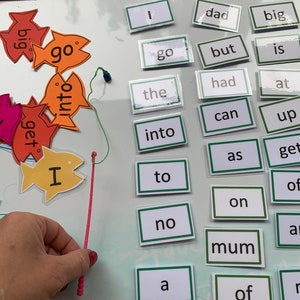May include: A white board with a fishing rod and colourful fish with words on them. The words on the fish include "go", "into", "get", and "I". There are also word cards on the board with words like "dad", "big", "go", "but", "is", "had", "at", "can", "up", "get", "as", "to", "no", "a", "mum", "of", and "r".
